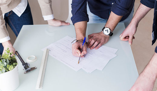 Tres personas revisando y firmando documentos sobre una mesa, con una planta y herramientas de medición visibles.