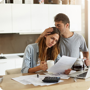 Mujer con gesto pensativo revisando documentos en una cocina, mientras un hombre a su lado la reconforta.