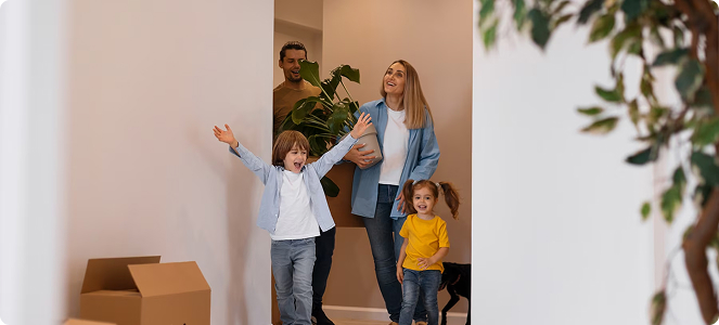 Familia con dos niños entrando a un nuevo hogar con cajas y una planta en las manos.
