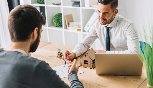 Hombre en traje señalando un documento sobre la mesa mientras otro hombre lo observa. En la mesa, hay una maqueta de casa, una laptop y un tarro con monedas.