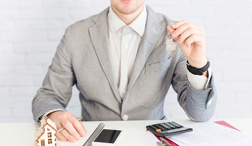 Hombre con traje sosteniendo unas llaves sobre un escritorio con documentos, calculadora y una maqueta de casa.