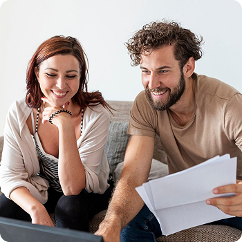 Pareja sonriendo mientras revisa documentos y usa un ordenador portátil, representando planificación financiera o gestión de presupuesto.