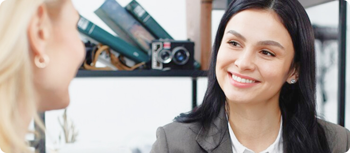 Asesora financiera sonriente conversando con una clienta en una oficina, con estantes y cámaras de decoración al fondo.
