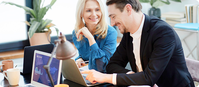Hombre y mujer en una oficina, sonriendo mientras consultan gráficas financieras en un portátil.