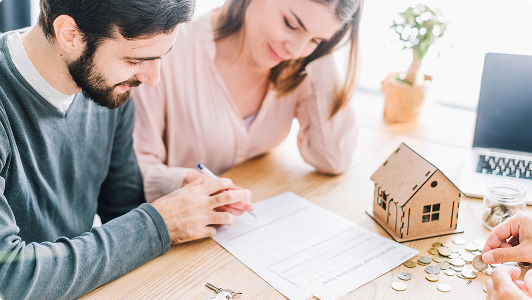 Pareja firmando un documento en una mesa con monedas, llaves y una maqueta de casa.