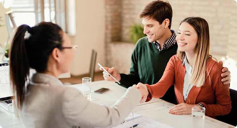 Pareja sonriente estrechando la mano de una asesora financiera en una oficina, con documentos y una tablet sobre la mesa.