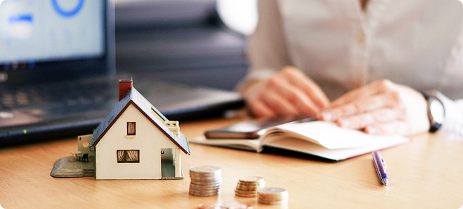 Maqueta de una casa sobre una mesa con monedas apiladas y una persona tomando notas junto a un ordenador portátil.