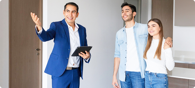 Agente inmobiliario mostrando un piso a una pareja joven, señalando con la mano mientras sostiene una tableta.