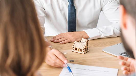Persona con traje sentado en una mesa junto a una maqueta de casa, monedas y un documento.