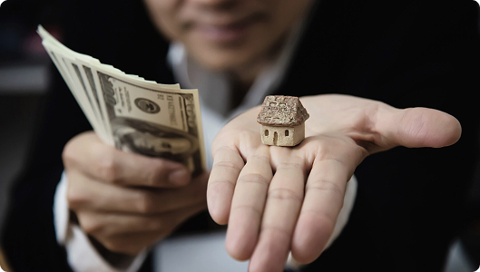 Persona sosteniendo una maqueta de casa en una mano y billetes en la otra, representando inversión en bienes raíces o financiamiento hipotecario.