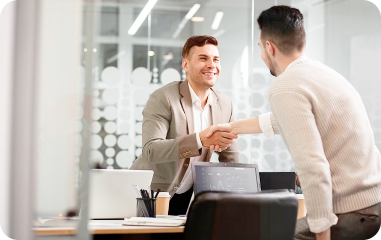 Asesor financiero estrechando la mano con un cliente en una oficina, con un portátil y documentos sobre la mesa.