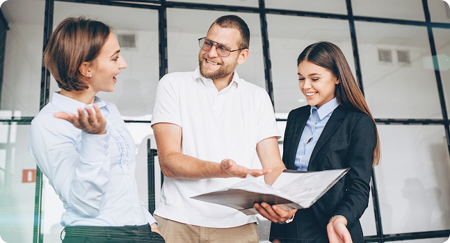 Grupo de tres personas conversando y revisando documentos en un entorno empresarial con ventanales.